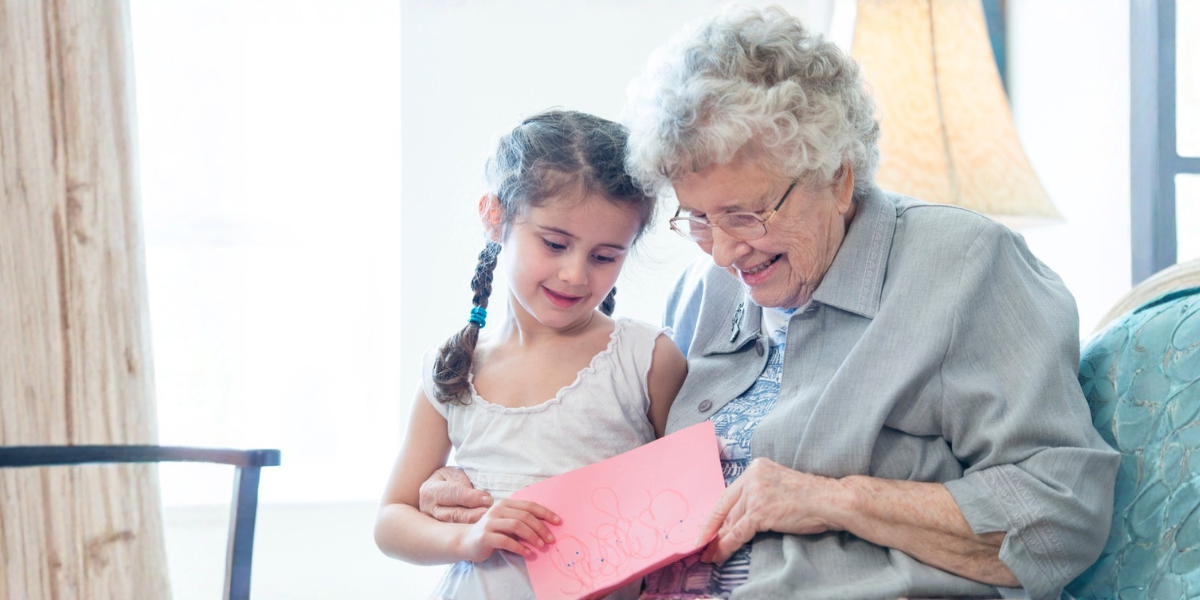 A little girl with braids is giving a card to an elderly woman. They are smiling